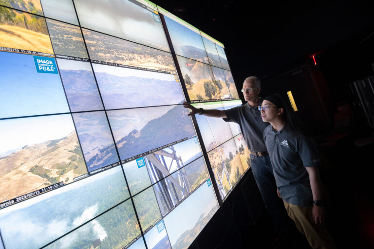Two University of California researchers gesture to a floor-to-ceiling monitor with several landscape feeds on display.