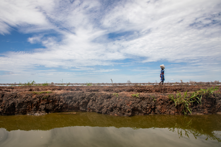 ICPAC’s early warning system project receives funding from CRAF’d A woman walks carrying a bag on her head, on a mud path next to a body of water.