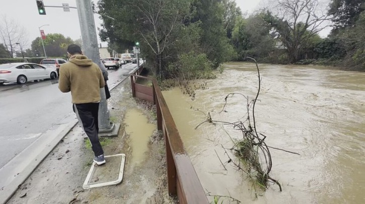 FEATURE: How did Stanford University and the University of Florida create a model to simulate urban flood risk with an eye to equity? A man walks along a road, past a muddy overflowing river that has a tree poking out.