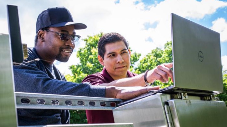 Virginia Tech researchers use deep learning to predict flooding Two men work cheerfully at a laptop, with a blue sky and trees in the background.