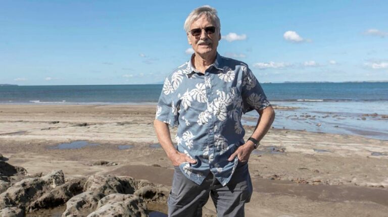 Dr Kevin Trenberth, a climate scientist a the University of Auckland in New Zealand, stands on a beach with his hands in his front pockets and the ocean behind him.