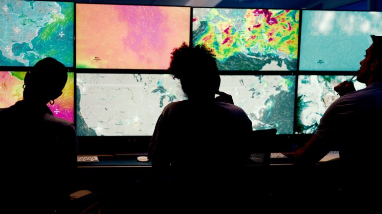 Three people sit in front of a wide bank of computer screens displaying meteorological maps and charts.