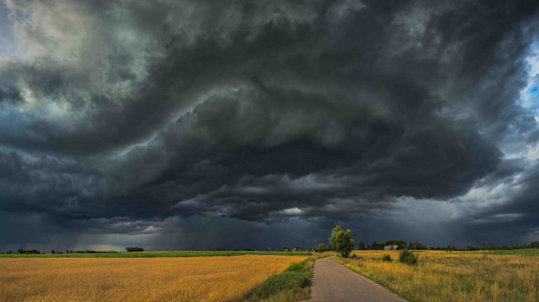 A large storm with black swirling clouds builds over a rural scene, with croops, sparse trees and a road that runs through the center of the image into the distance.